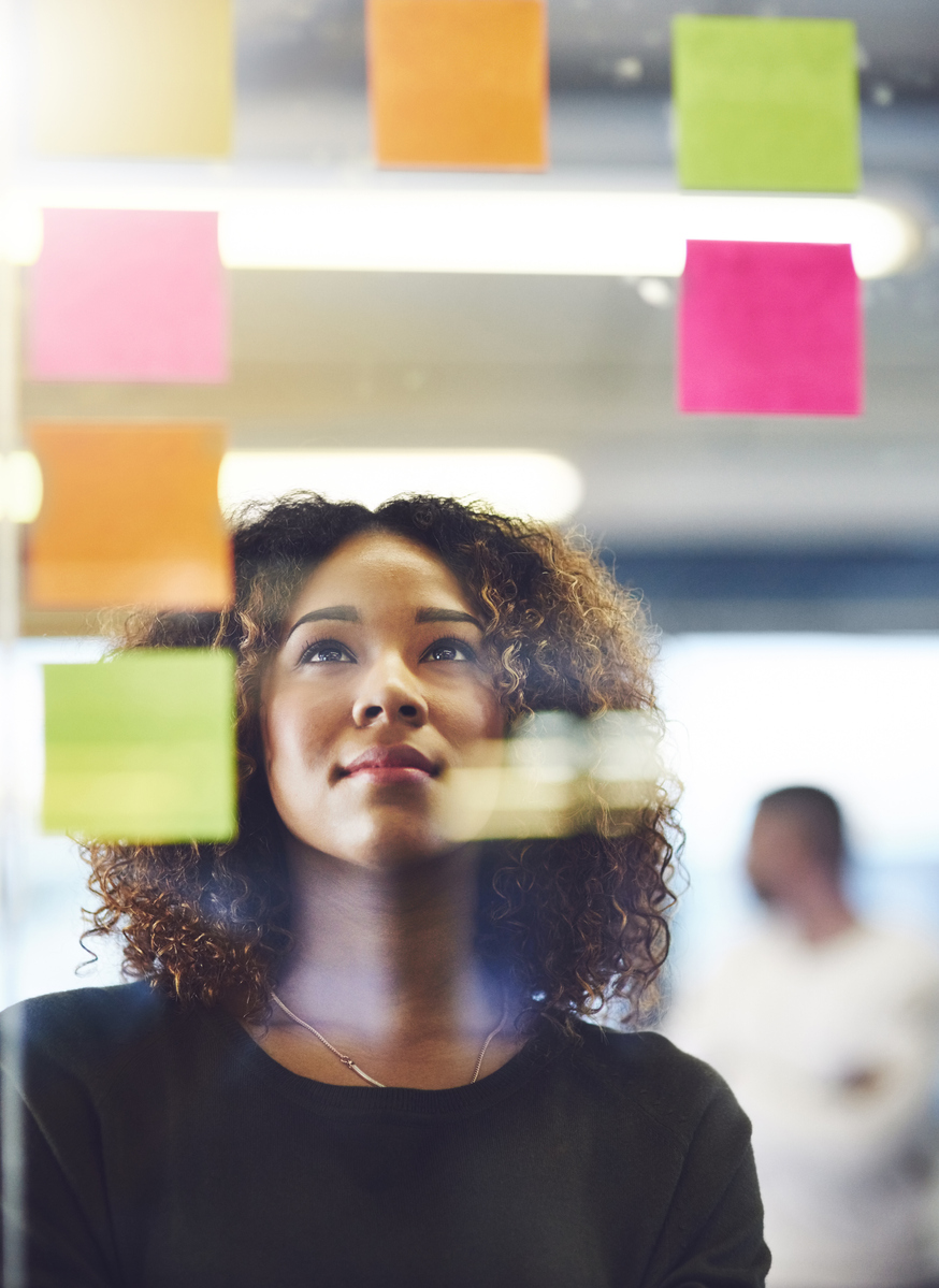 Junge Frau steht vor Kanban-Board mit bunten Post-its und überlegt die nächsten Schritte.