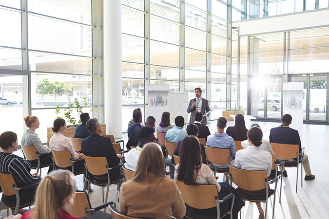 Personengruppe sitzt in einem modernen, hellen Konferenzraum und hört einem Vortrag zu; vorne steht eine Person neben einem beschrifteten Whiteboard.