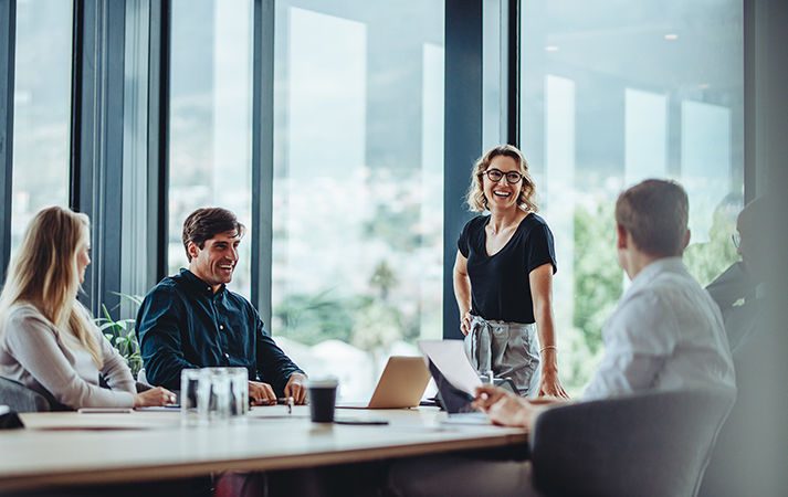 Vier Personen in einem modernen Büro bei einer Besprechung; eine Person steht und spricht, die anderen sitzen am Tisch mit Laptops und Unterlagen.