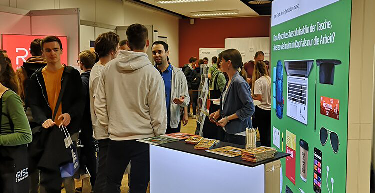 Personengruppe an einem Messestand mit grünem Banner, das zur Berufsorientierung nach der Matura auffordert; auf dem Tisch liegen Broschüren und Werbematerialien.