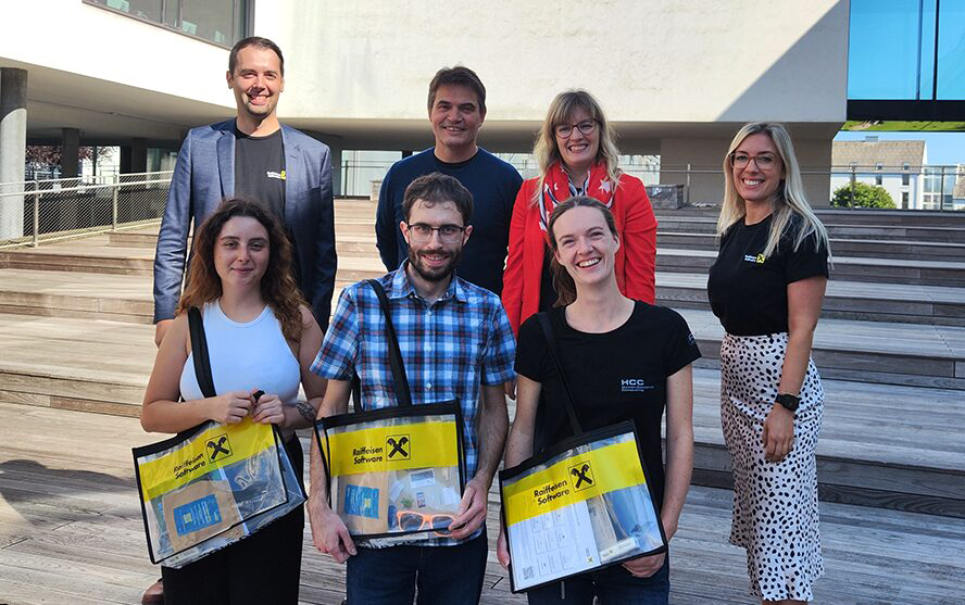 Sieben Personen stehen auf Holzstufen vor einem modernen Gebäude; drei von ihnen halten transparente Taschen mit ‚Raiffeisen Software‘-Branding.