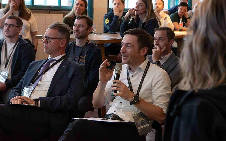 Teilnehmer:innen sitzen in einem Konferenzraum; eine Person im Vordergrund spricht ins Mikrofon – Szene aus einer interaktiven Veranstaltung.