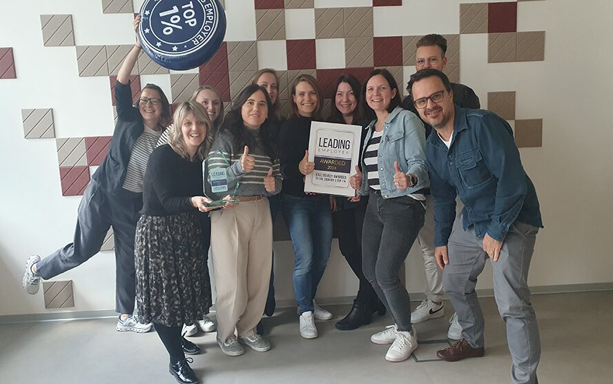Neun Personen posieren mit Auszeichnungen vor einer Wand mit geometrischem Muster; darunter ein Schild mit ‚Top 1% Employer‘ und eine Plakette mit ‚Leading Employer Awarded‘.