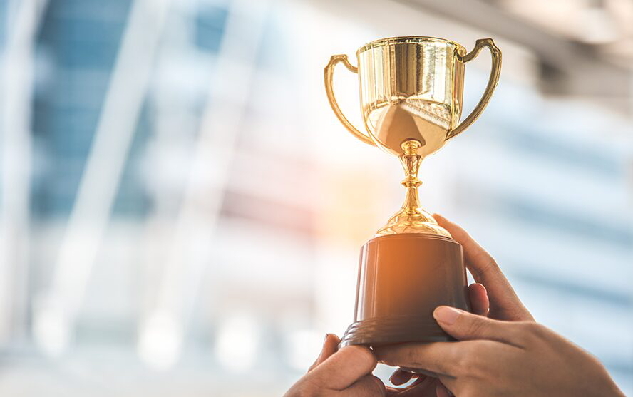 Mehrere Hände halten gemeinsam einen goldenen Pokal mit Henkeln – Symbol für Erfolg und Auszeichnung.