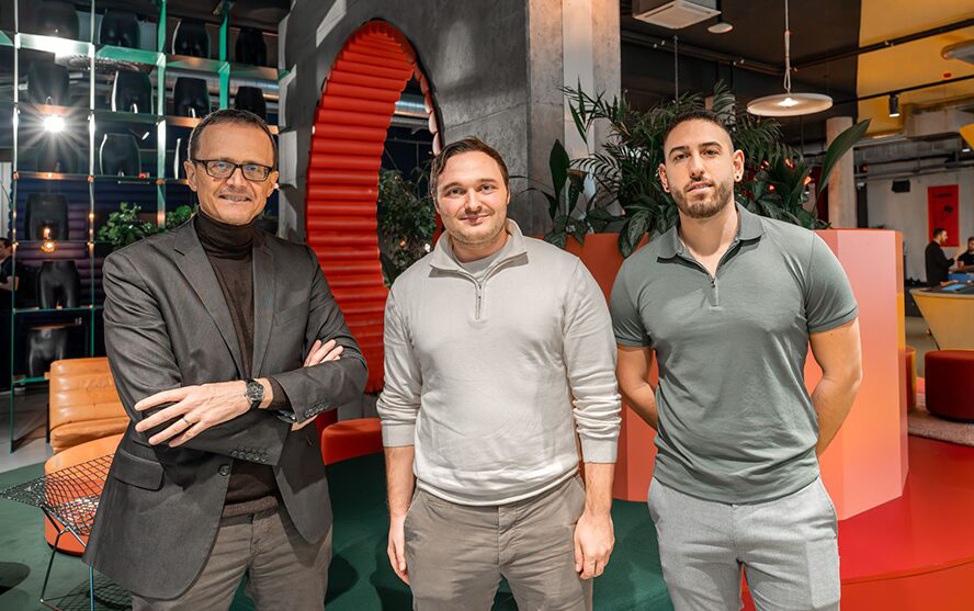 Drei Personen stehen in einem modern gestalteten Innenraum mit bunten Möbeln, Pflanzen und einer roten Treppe im Hintergrund.