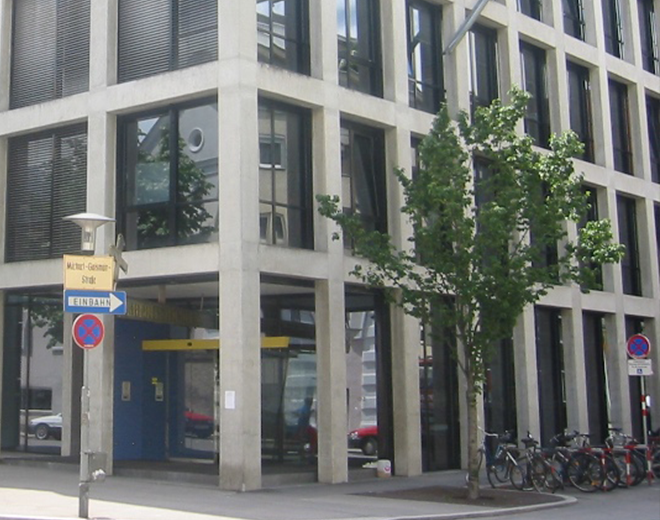 Modernes Gebäude mit großen Fenstern und Betonrahmen; vor dem Eingang mit blauer Tür stehen Fahrräder, ein Baum und Straßenschilder mit ‚Einbahn‘ und ‚Münchener Straße‘.