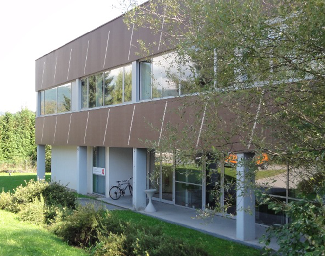 Modernes zweistöckiges Gebäude mit großen Fenstern und brauner Fassade, umgeben von Grünflächen; ein Fahrrad steht nahe dem Eingang.