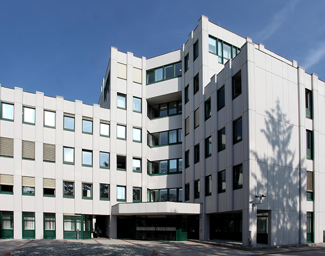 Modernes Bürogebäude mit weißer Fassade und markanter Architektur vor blauem Himmel – Schatten eines Baumes fällt auf die rechte Seite.