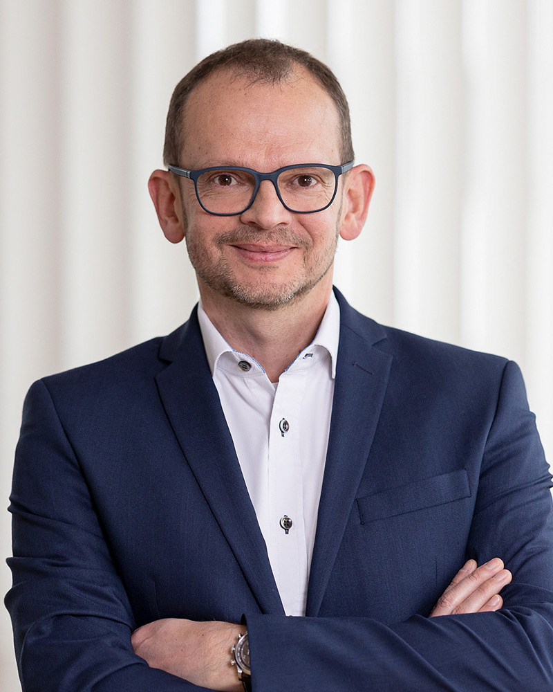 Porträt eines Mannes mit Brille, Arme verschränkt, blaues Sakko.