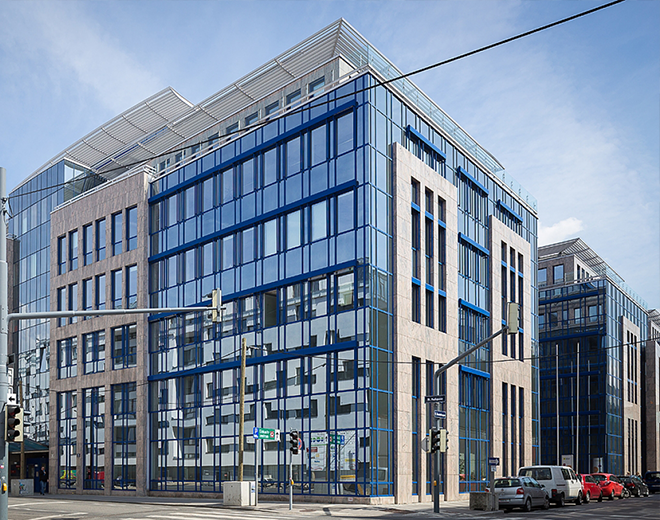 Mehrstöckiges Bürogebäude mit Glasfassade und blauen Akzenten an einer Straßenkreuzung; Autos und Verkehrsschilder im Vordergrund.