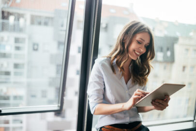 Person sitzt an einem großen Fenster mit Blick auf städtische Gebäude und nutzt ein Tablet.