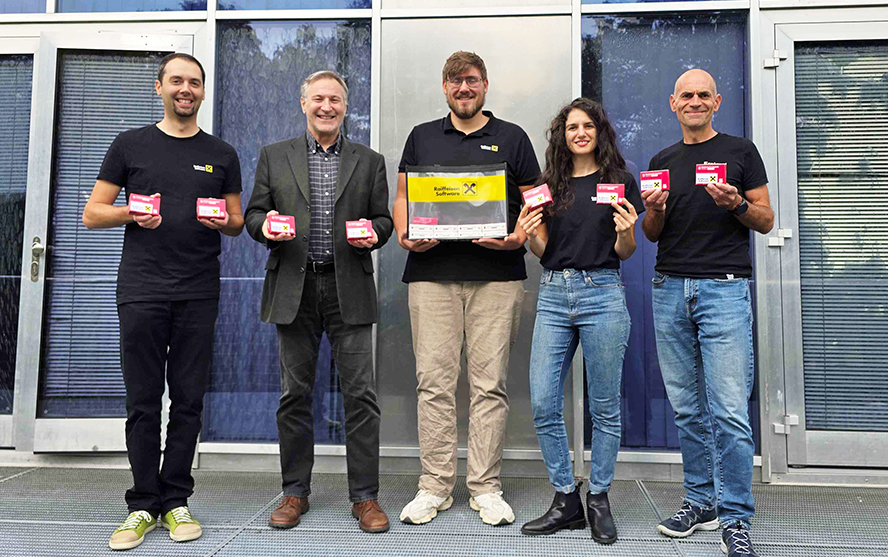 Fünf Personen stehen vor einem modernen Gebäude und halten mehrere kleine rote Karten sowie eine schwarze Tasche mit Raiffeisen Software Logo.