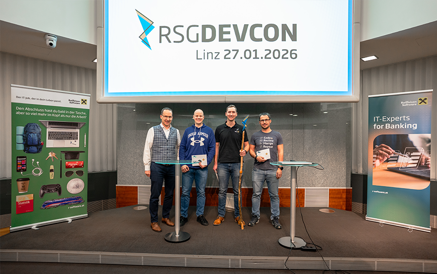 Vier Personen stehen auf einer Bühne vor einer großen Leinwand mit dem Logo ‚RSG DEVCON‘ und dem Datum ‚Linz 27.01.2026‘. Zwei Roll‑up‑Banner flankieren die Bühne: links einer mit Werbegeschenken und Raiffeisen‑Branding, rechts einer mit der Aufschrift ‚IT‑Experts for Banking‘. Die Personen halten Urkunden und Preise, darunter zwei große Wanderstöcke. Die Szene wirkt wie eine Preisverleihung oder Ehrung im Rahmen der Veranstaltung.