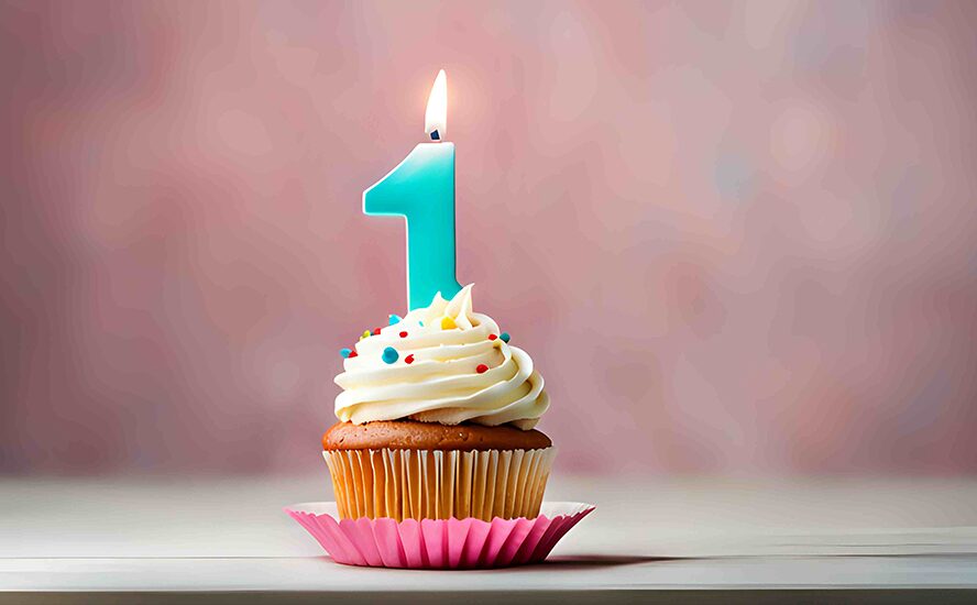 Cupcake mit weißem Frosting und bunten Streuseln, darauf eine brennende blaue Kerze in Form der Zahl 1 vor unscharfem Hintergrund.