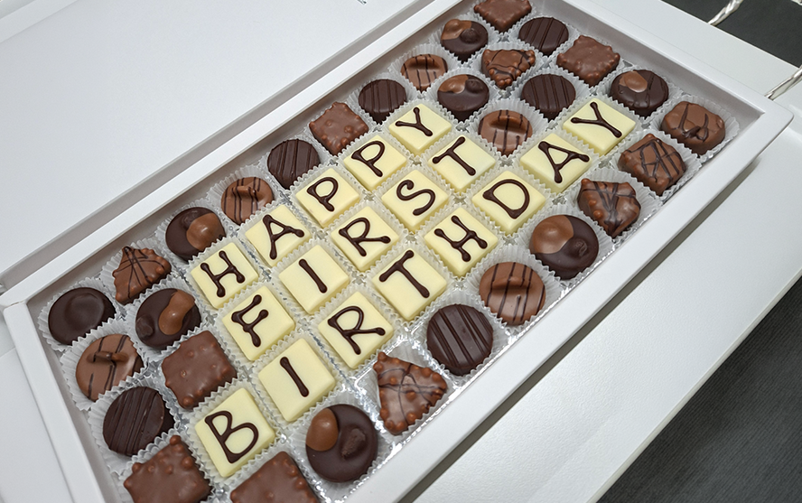 Geöffnete Schachtel mit Pralinen; in der Mitte stehen aus weißen Schokoladenwürfeln geformte Buchstaben, die den Text Happy First Birthday bilden. Rundherum sind verschiedene dunkle und helle Pralinen dekorativ angeordnet.