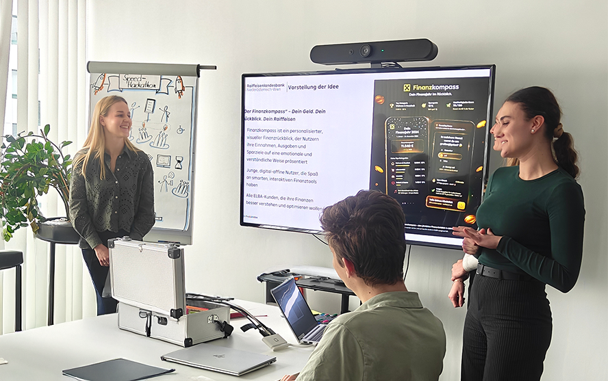 Drei Personen in einem Meetingraum während einer Präsentation mit Bildschirm, Laptop und Flipchart.